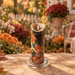 May include: A cylindrical pillar candle with a black background featuring a colorful floral pattern of orange, red, and purple flowers. The candle sits on a clear glass circular plate and has a small, lit flame at the top center of the wax.