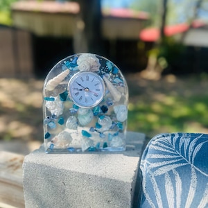 May include: A clear resin clock with a silver clock face and Roman numerals. The clock is filled with seashells and blue and white stones. The clock is sitting on a gray concrete surface.