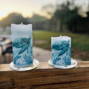 May include: Two battery-operated, cylindrical candles with a sea turtle design. The candles are white with a blue and teal turtle illustration and sit on clear glass saucers. The background is blurred, with a wooden railing in the foreground.