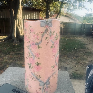 May include: A pink candle with a floral and ribbon design. The candle is sitting on a gray surface with a remote control in the foreground.