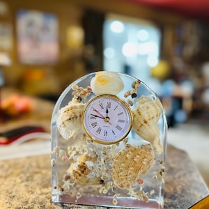 May include: A clear resin clock with a gold-tone bezel, featuring a white clock face with Roman numerals. The clock is adorned with various seashells and small pebbles. The clock is approximately 5 inches tall and 4 inches wide.