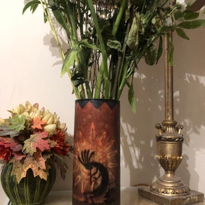 May include: A cylindrical vase featuring a dark brown and orange gradient finish with a black silhouette of a Kokopelli figure playing a flute. The top rim has a black zigzag pattern, and the base is a solid dark brown color.