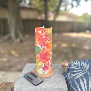 May include: A cylindrical, battery-operated candle with a fall-themed design. The candle features colorful leaves, pumpkins, and berries. A remote control is placed next to the candle. The candle is approximately 6 inches tall.