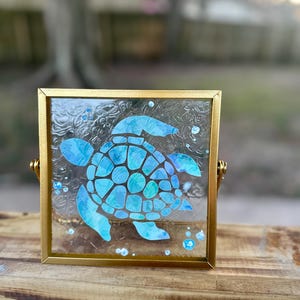 May include: A framed glass art piece with a blue and green sea turtle silhouette. The frame is gold and has a textured glass surface.