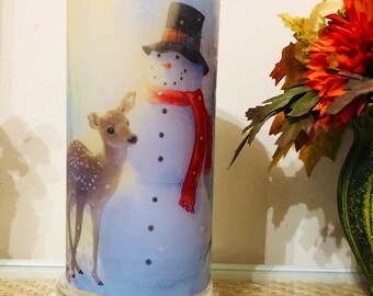 May include: A cylindrical glass candle holder with a winter scene featuring a snowman wearing a red scarf and a black top hat, and a deer standing next to him. The candle holder has a clear glass base with a beaded edge.
