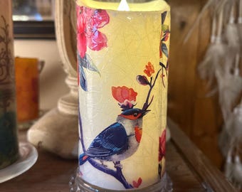 May include: A white pillar candle with a floral and bird design. The candle has a warm glow and is sitting on a clear glass base with a beaded edge.