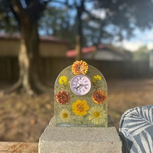 May include: A decorative clock with a floral design. The clock face is round with Roman numerals. The clock is surrounded by yellow, orange, and white flowers encased in clear resin. The clock is sitting on a gray concrete surface.