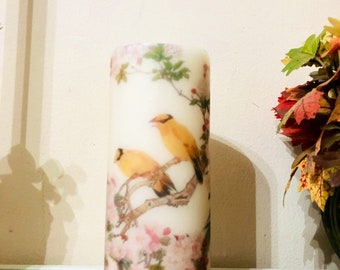 May include: A white pillar candle with a floral and bird design. The candle has a clear glass base with a beaded edge.
