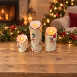 May include: Three cylindrical LED candles of varying heights featuring a cream-colored exterior with a printed pattern of red flowers, green pine needles, and brown foliage. Each candle has a glowing yellow flame-shaped light at the top.