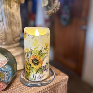 Könnte beinhalten: Eine batteriebetriebene, zylindrische Kerze mit Sonnenblumenmuster. Die Kerze hat einen gelb-weißen Hintergrund mit gelben Sonnenblumen, grünen Blättern und lila Blumen. Sie steht auf einer durchsichtigen Glasplatte und hat ein flackerndes LED-Licht.