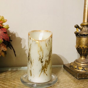 May include: A white pillar candle with gold leaf accents and a flickering LED flame. The candle is sitting on a clear glass base.