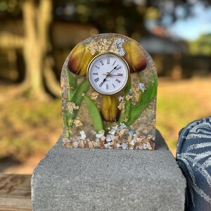 May include: A decorative clock with a gold-tone bezel and a white face with black Roman numerals. The clock is set within a clear resin block, featuring yellow and brown floral elements, green leaves, and gold flakes. The base is adorned with small seashells.