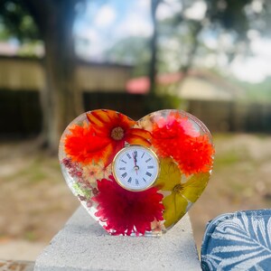 May include: A heart-shaped clock with a clear resin body, embedded with vibrant red, orange, and yellow flowers. The clock face is white with gold hands and Roman numerals. The clock is sitting on a gray surface.