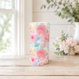May include: A cylindrical tumbler featuring a watercolor floral pattern with soft pink, peach, and light blue flowers and green leaves. The design covers the entire exterior surface of the vessel, showcasing a repeating botanical motif in pastel tones.