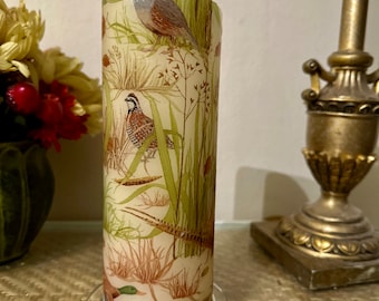 May include: A cylindrical candle with a white base and a colorful design featuring birds and foliage. The candle is on a clear glass base.