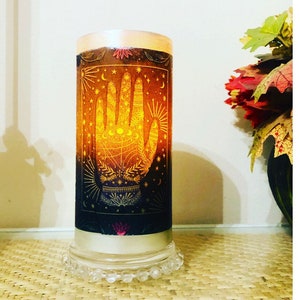 May include: A cylindrical candle with a palmistry design. The candle features a glowing hand illustration with stars and celestial symbols in shades of gold and black. The candle sits on a clear, beaded base, creating a mystical ambiance.