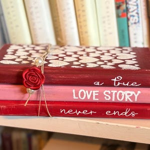 May include: A stack of three decorative books painted in shades of red and pink. The top book is burgundy with white heart designs. The middle book is pink, and the bottom book is red. Text on the books reads "a true LOVE STORY never ends."