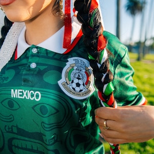 May include: Green Mexico soccer jersey with white collar and the word "MEXICO" on the front. The jersey features a detailed design and a team emblem. Braided hair is adorned with red, green, and white ribbons, reflecting the colors of the Mexican flag.