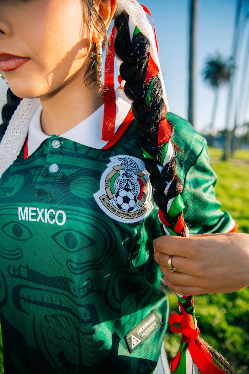 May include: Green Mexico soccer jersey with white collar and the word "MEXICO" on the front. The jersey features a detailed design and a team emblem. Braided hair is adorned with red, green, and white ribbons, reflecting the colors of the Mexican flag.