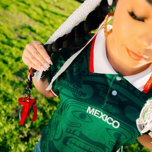 May include: Green Mexico jersey with white collar and red trim. The jersey features a detailed pattern and the word "MEXICO" in white. The image also shows a braided hairstyle with red, white, and green ribbons.