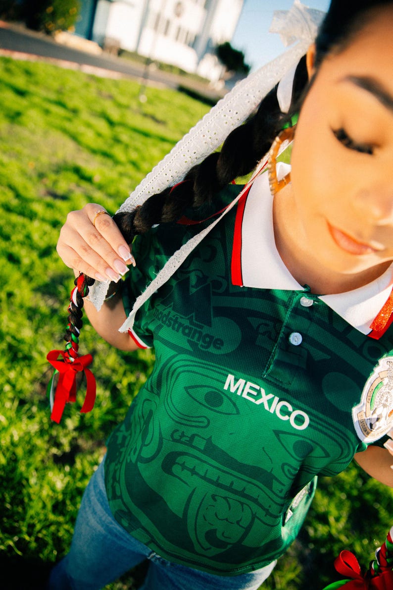 May include: Green Mexico jersey with white collar and red trim. The jersey features a detailed pattern and the word "MEXICO" in white. The image also shows a braided hairstyle with red, white, and green ribbons.