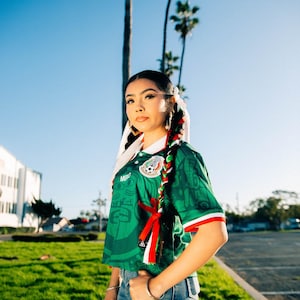 May include: A person wearing a green soccer jersey with red and white accents, and the Mexican flag emblem. The jersey is paired with blue jeans. The background includes palm trees and a clear blue sky.