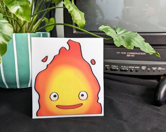 May include: A white card with a cartoon drawing of a yellow and orange fire spirit with a smiling face and black outlines. The fire spirit has two large eyes and a small mouth.