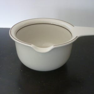 May include: A white ceramic gravy boat with a handle and a spout. The rim and handle are outlined with a thin brown line. The gravy boat is on a dark surface.