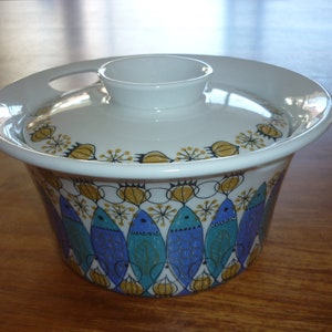Può includere: Piatto da portata in ceramica bianca con coperchio. Il piatto presenta un motivo a pesci blu, verdi e dorati. Il coperchio ha un motivo floreale dorato e bianco.