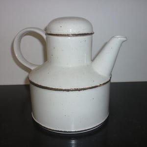Op de afbeelding: Een witte keramische theepot met een deksel, handvat en tuit. De theepot heeft een cilindrische basis en een afgeronde bovenkant. Hij is bespikkeld met kleine bruine stippen en heeft bruine randen.