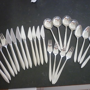 Könnte beinhalten: Eine Sammlung glänzender, silberfarbener Besteckteile, darunter Gabeln, Messer und Löffel, angeordnet auf einer dunklen Oberfläche. Das Besteck hat ein schlichtes, elegantes Design und reflektiert das Licht. Das Set enthält verschiedene Löffelgrößen.