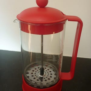 May include: A French press coffee maker featuring a clear glass carafe, a bright red plastic lid with a round knob, a matching red plastic base, and a red side handle. Inside the carafe is a metal plunger assembly with a circular filter screen.