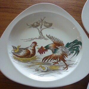 May include: A white oval ceramic plate with a raised rim features a colourful illustration of a rooster, hen, and chicks. The rooster is brown, red, and green, while the hen is brown and the chicks are yellow. The plate has a rustic farmhouse theme.