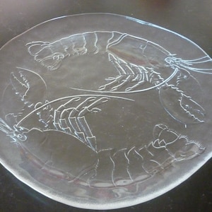 May include: Clear glass plate with etched lobster design. The plate features two detailed lobster illustrations, showcasing their claws, bodies, and antennae. A decorative serving dish for seafood or coastal-themed dining.