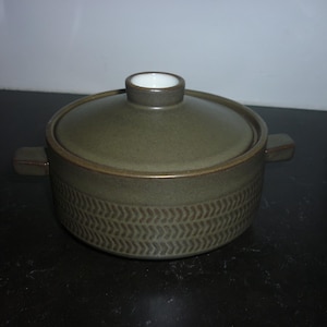 May include: An olive green ceramic casserole dish with a lid. The dish features a textured pattern around the base and a brown rim. It has two small handles and a white interior on the lid's knob.