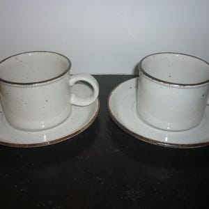 Op de afbeelding: Twee crèmekleurige keramische koffiekopjes met bijpassende schotels. Elk kopje heeft een oor en een donkerbruine rand. De schotels hebben ook een donkerbruine rand. De kopjes en schotels hebben een gespikkeld ontwerp.