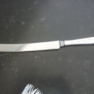 May include: A silver bread knife with a serrated edge and a decorative handle. The handle has a floral design.