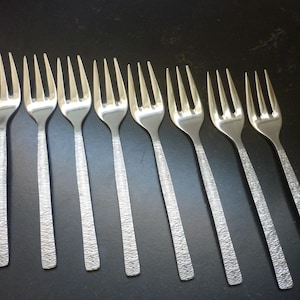 May include: A set of eight stainless steel forks arranged in a row. Each fork features a polished three-tine head and a flat, rectangular handle with a distinct, textured, cross-hatched finish that runs vertically along the length of the handle.