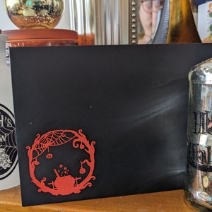 Può includere: Un biglietto nero con un design rosso a tema Halloween con un calderone, ragnatele e zucche. Sono visibili anche una bottiglia bianca con il testo "Witch's Brew" e una bottiglia di vetro decorativa con il testo "Happy Halloween".