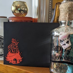 May include: A black card with a red illustration of a cauldron with steam rising from it. The card is sitting on a wooden surface with a clear glass bottle with a white charm and the text "Happy Halloween" on it.