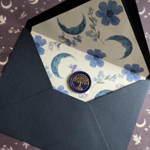 May include: A dark blue envelope with a white inner liner featuring a watercolour design of blue moons and flowers. A gold wax seal with a tree design is on the front of the envelope.