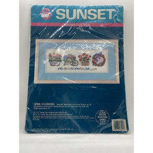 Op de afbeelding: Sunset geteld kruissteekpatroon met een kleurrijke opstelling van lentebloemen in manden en een kruiwagen. Het patroon is getiteld "April Flowers" en bevat instructies voor een framemaat van 40x15cm zonder passe-partout of een framemaat van 50x25cm met passe-partout.