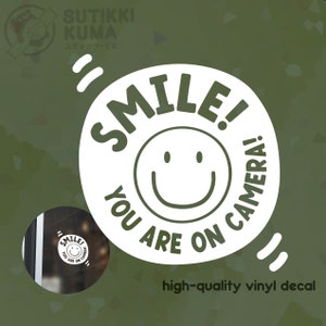 May include: White vinyl decal with the text "SMILE! YOU ARE ON CAMERA!" in a circular design, featuring a smiling face. The decal also includes the text "high-quality vinyl decal".