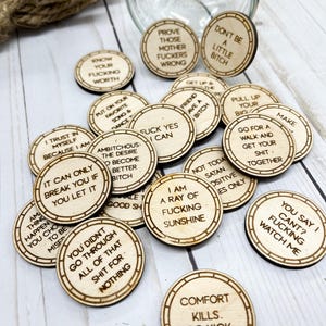 May include: A collection of round wooden tokens with various motivational and humorous phrases laser-etched onto them. The tokens are light brown with dark brown borders and text. Some phrases include "Know Your Fucking Worth" and "Don't Be a Little Bitch."
