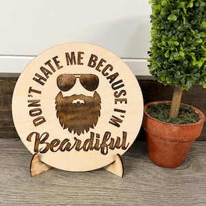 May include: A round wooden sign with the text "DON'T HATE ME BECAUSE I'M Beardiful" and a graphic of a bearded face wearing sunglasses. The sign is displayed on a wooden stand. A small potted plant is to the right.