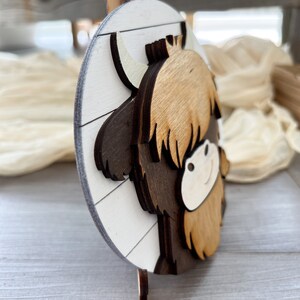 May include: A wooden, layered Highland cow decoration. The cow features a light brown shaggy coat, dark brown horns, and a white face. The background is a white circle with wooden slats. The decoration stands on a small wooden base.