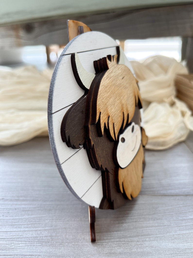 May include: A wooden, layered Highland cow decoration. The cow features a light brown shaggy coat, dark brown horns, and a white face. The background is a white circle with wooden slats. The decoration stands on a small wooden base.