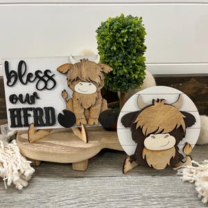 May include: Wooden Highland cow decor with a sign that reads "bless our herd". The sign and cow are on a wooden stand with a small green plant. The cow is brown and tan with a smiling face.
