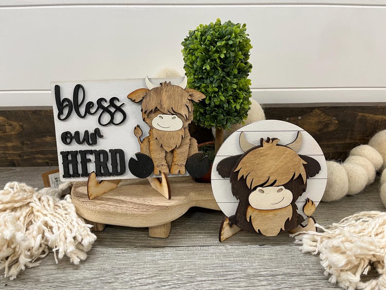 May include: Wooden Highland cow decor with a sign that reads "bless our herd". The sign and cow are on a wooden stand with a small green plant. The cow is brown and tan with a smiling face.