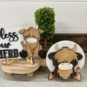 May include: Wooden farmhouse decor featuring a Highland cow and a sign that reads "bless our herd." The sign is white with black lettering. The cow figurines are brown and tan, with a smiling face. A small green plant is also included.